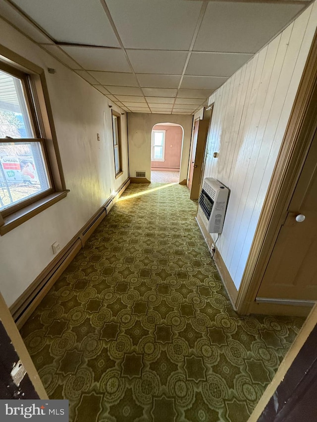 hallway with a paneled ceiling, dark carpet, heating unit, wooden walls, and a baseboard radiator