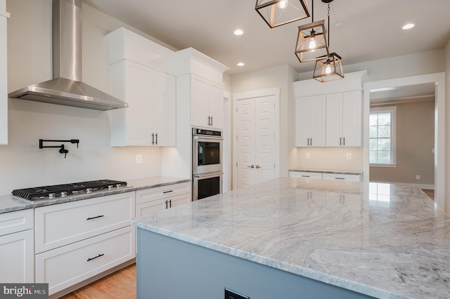 kitchen with hanging light fixtures, wall chimney range hood, stainless steel appliances, light stone countertops, and white cabinets