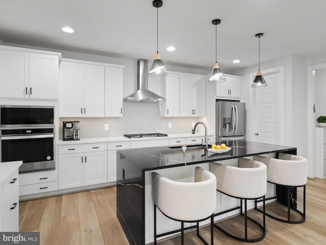 kitchen with sink, wall chimney exhaust hood, an island with sink, appliances with stainless steel finishes, and white cabinetry