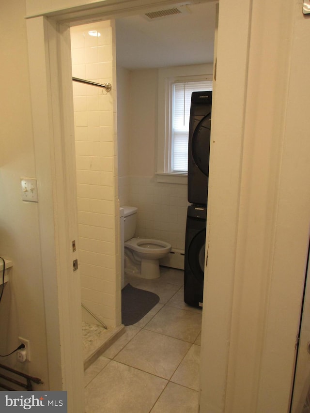 bathroom featuring tile patterned flooring, toilet, stacked washing maching and dryer, and tile walls