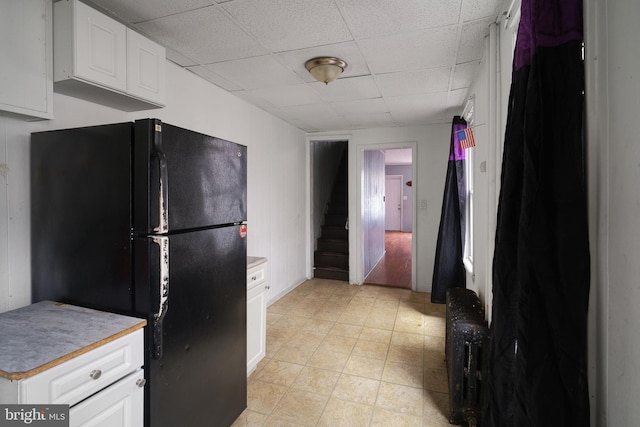 kitchen with white cabinetry, a drop ceiling, radiator heating unit, black refrigerator, and light tile patterned floors