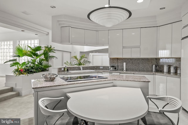 kitchen with decorative backsplash, light stone countertops, black electric stovetop, white cabinets, and hanging light fixtures