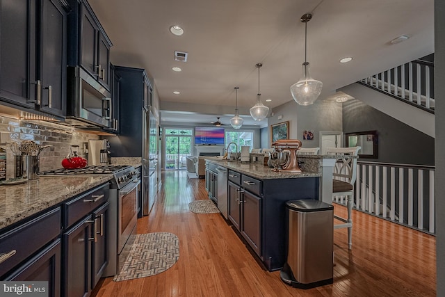 kitchen with a kitchen breakfast bar, appliances with stainless steel finishes, light stone counters, a center island with sink, and hanging light fixtures