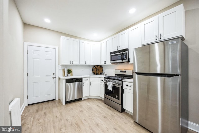 kitchen featuring light wood-type flooring, backsplash, radiator, stainless steel appliances, and white cabinetry