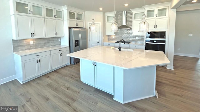 kitchen with tasteful backsplash, wall chimney range hood, a center island with sink, light stone countertops, and stainless steel appliances