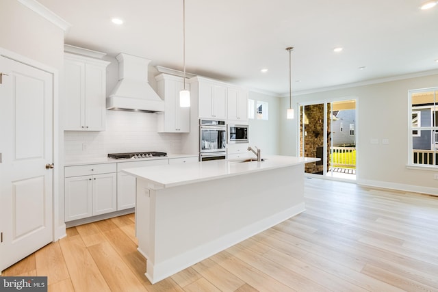 kitchen featuring appliances with stainless steel finishes, premium range hood, white cabinetry, hanging light fixtures, and an island with sink