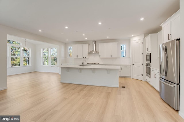 kitchen featuring a center island with sink, white cabinets, wall chimney range hood, appliances with stainless steel finishes, and light hardwood / wood-style floors