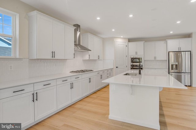 kitchen with light wood-type flooring, wall chimney exhaust hood, stainless steel appliances, sink, and a center island with sink