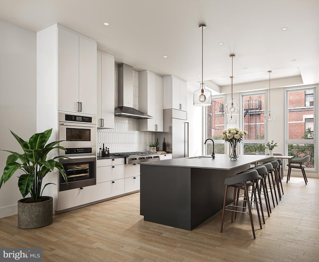 kitchen featuring white cabinetry, wall chimney exhaust hood, hanging light fixtures, an island with sink, and light hardwood / wood-style floors