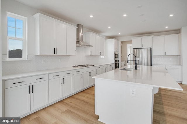 kitchen with sink, wall chimney exhaust hood, stainless steel appliances, an island with sink, and white cabinets