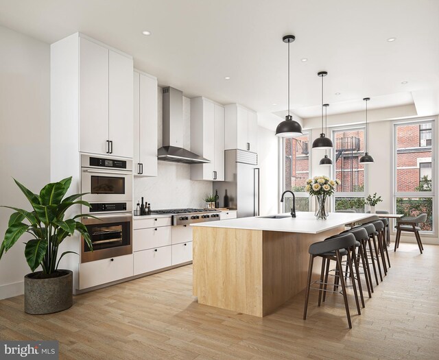 kitchen featuring white cabinets, light wood-type flooring, wall chimney exhaust hood, and an island with sink