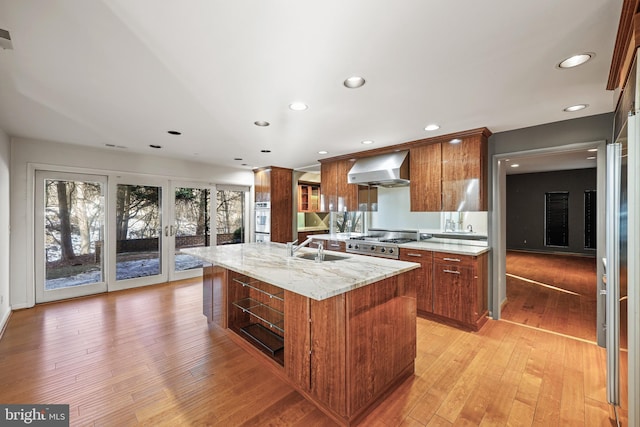 kitchen featuring wall chimney range hood, appliances with stainless steel finishes, light stone countertops, a center island with sink, and light wood-type flooring