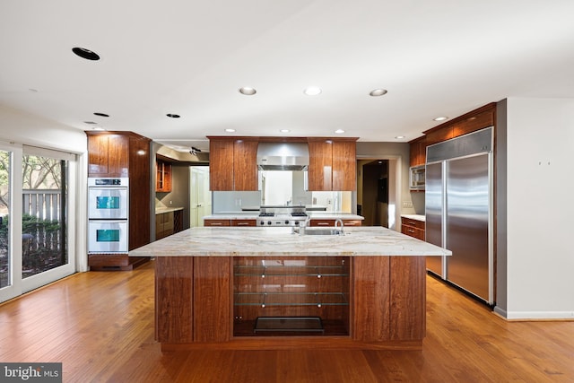 kitchen with appliances with stainless steel finishes, a center island, wall chimney exhaust hood, light stone counters, and light hardwood / wood-style flooring