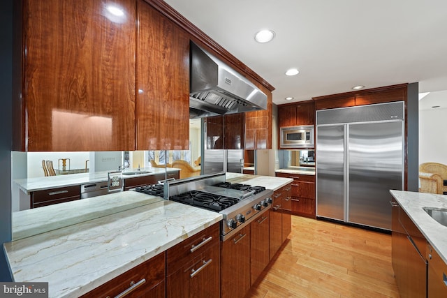 kitchen featuring wall chimney exhaust hood, sink, built in appliances, light wood-type flooring, and light stone countertops