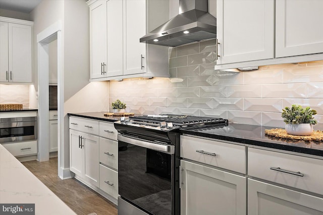 kitchen with appliances with stainless steel finishes, wall chimney exhaust hood, dark stone counters, and white cabinetry