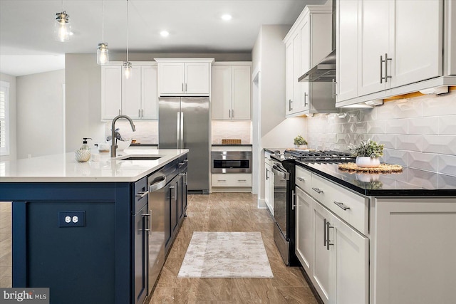 kitchen featuring wall chimney range hood, stainless steel appliances, sink, hanging light fixtures, and a kitchen island with sink