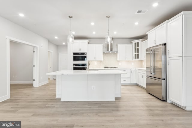 kitchen featuring wall chimney exhaust hood, hanging light fixtures, a center island with sink, white cabinets, and appliances with stainless steel finishes