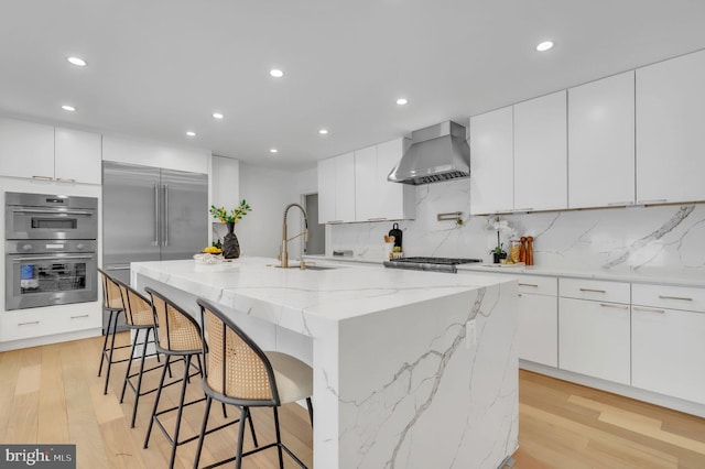 kitchen featuring wall chimney range hood, a center island with sink, sink, stainless steel appliances, and white cabinets