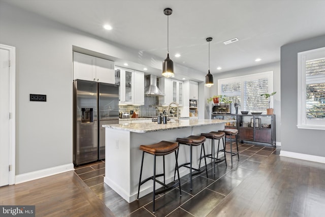 kitchen featuring stainless steel refrigerator with ice dispenser, wall chimney exhaust hood, dark wood-type flooring, pendant lighting, and white cabinetry