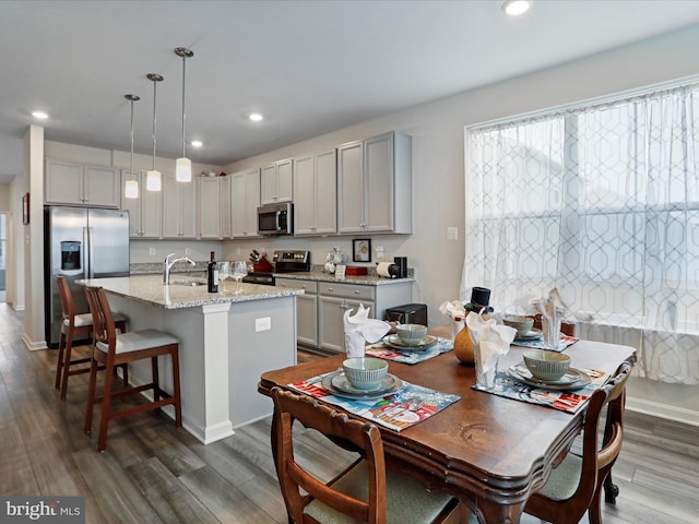 kitchen with light stone countertops, dark wood-type flooring, hanging light fixtures, stainless steel appliances, and a kitchen island with sink