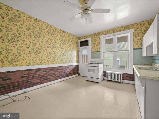 kitchen with radiator, sink, white gas stove, white cabinetry, and brick wall