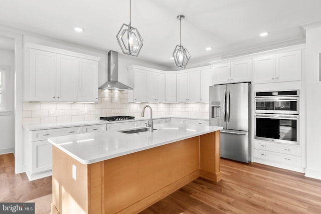 kitchen featuring sink, wall chimney exhaust hood, stainless steel appliances, a center island with sink, and white cabinets