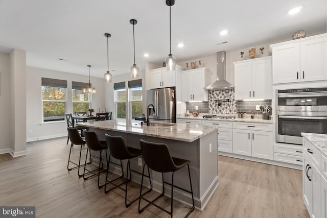 kitchen with white cabinets, decorative light fixtures, wall chimney range hood, and an island with sink