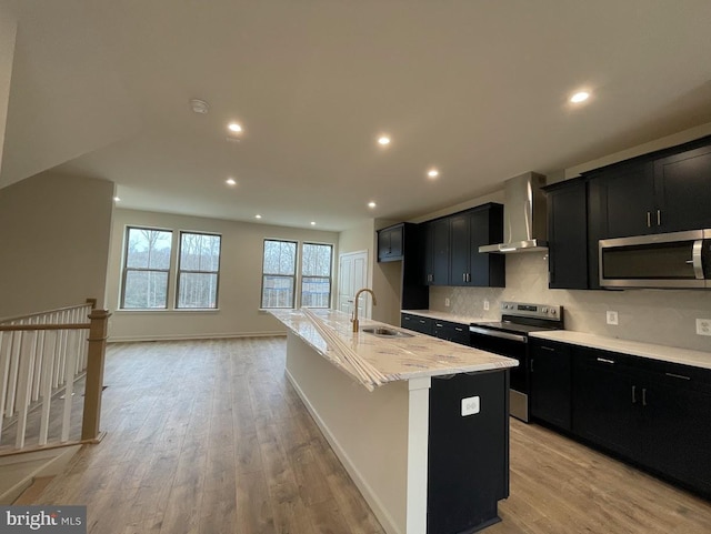 kitchen featuring appliances with stainless steel finishes, light wood-type flooring, a kitchen island with sink, sink, and wall chimney range hood