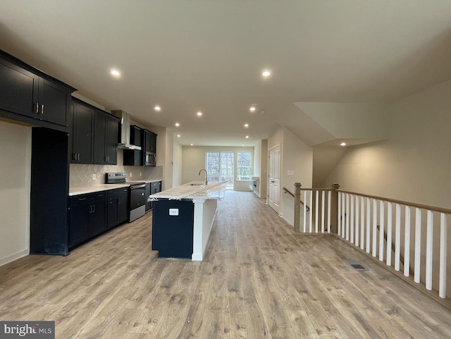 kitchen with wall chimney range hood, tasteful backsplash, light hardwood / wood-style floors, a kitchen island with sink, and appliances with stainless steel finishes