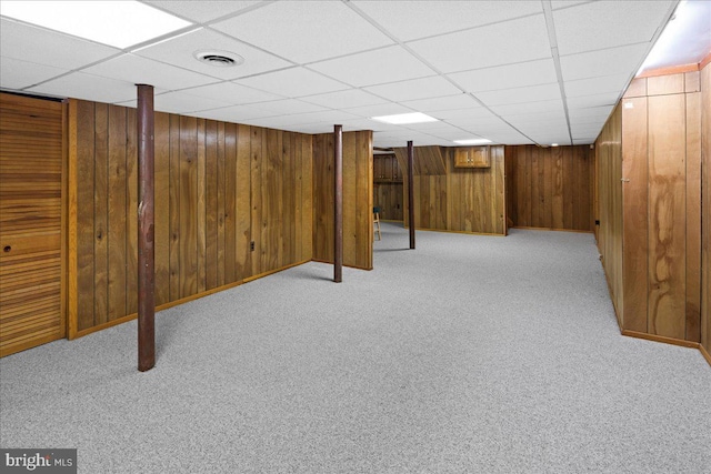 finished below grade area with a drop ceiling, visible vents, light carpet, and wood walls