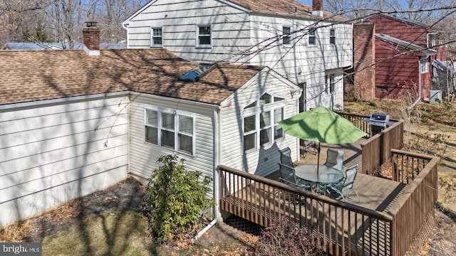 back of property with a deck, outdoor dining space, roof with shingles, and a chimney