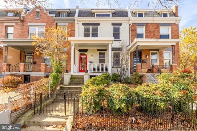 multi unit property featuring brick siding, a fenced front yard, roof with shingles, covered porch, and mansard roof