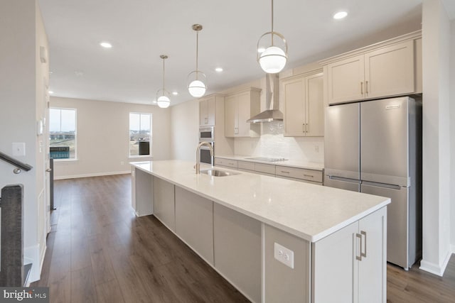 kitchen featuring wall chimney exhaust hood, stainless steel fridge, an island with sink, and decorative light fixtures