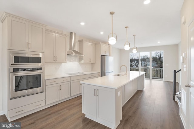 kitchen featuring appliances with stainless steel finishes, a kitchen island with sink, sink, wall chimney range hood, and pendant lighting