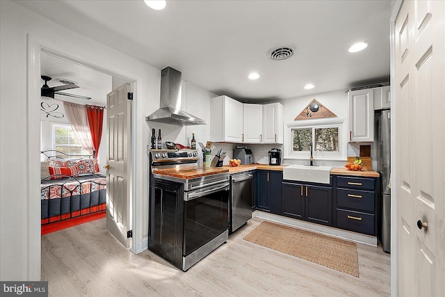 kitchen with stainless steel appliances, wall chimney range hood, sink, white cabinets, and butcher block countertops