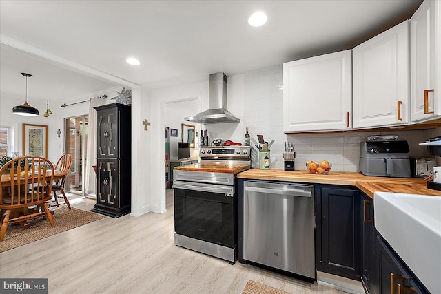 kitchen with wood counters, decorative backsplash, stainless steel appliances, wall chimney range hood, and hanging light fixtures