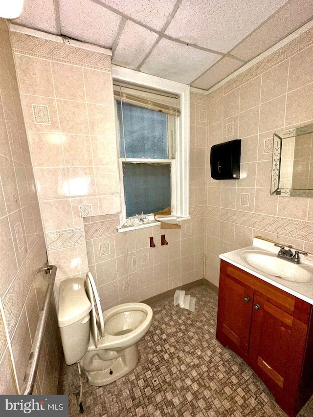 bathroom with a drop ceiling, vanity, tile walls, and toilet