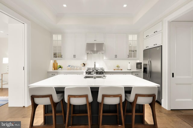 kitchen featuring wall chimney exhaust hood, stainless steel appliances, a tray ceiling, sink, and white cabinetry