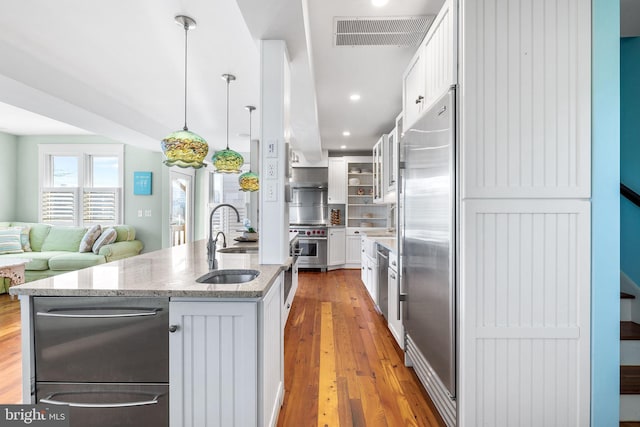 kitchen with light stone counters, pendant lighting, a center island with sink, built in fridge, and white cabinetry