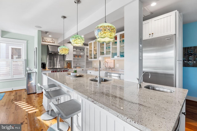 kitchen featuring a kitchen island with sink, stainless steel appliances, wall chimney exhaust hood, white cabinetry, and decorative light fixtures