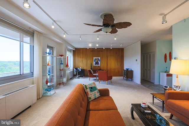 carpeted living room with wooden walls, radiator heating unit, ceiling fan, and rail lighting