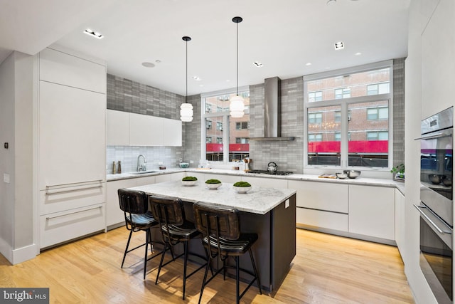 kitchen featuring white cabinetry, light wood-type flooring, stainless steel gas cooktop, wall chimney range hood, and a center island