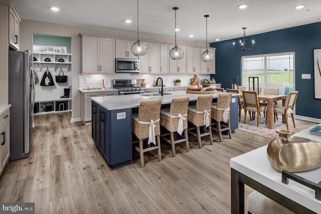 kitchen with sink, an island with sink, pendant lighting, appliances with stainless steel finishes, and light wood-type flooring