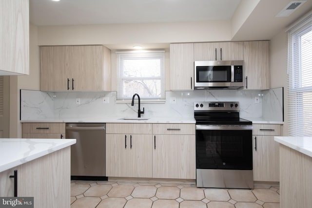 kitchen with light stone countertops, sink, backsplash, light brown cabinetry, and appliances with stainless steel finishes