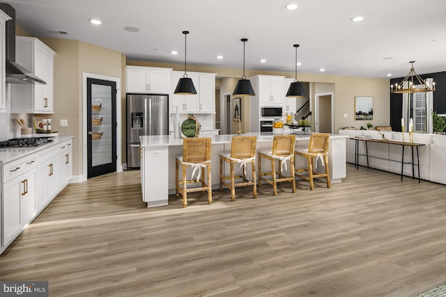 kitchen featuring white cabinetry, hanging light fixtures, stainless steel appliances, wall chimney range hood, and an island with sink