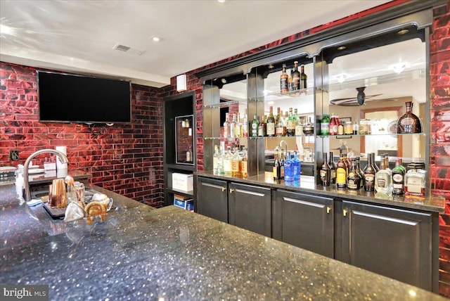 bar featuring ceiling fan, sink, dark stone counters, and brick wall