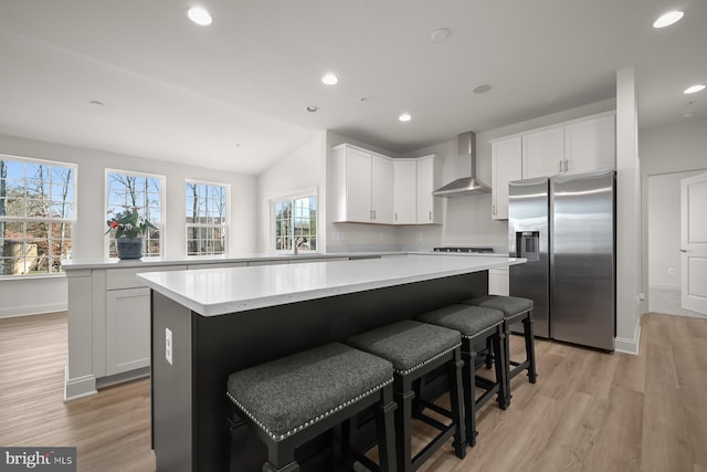 kitchen featuring light wood-type flooring, wall chimney exhaust hood, white cabinetry, stainless steel fridge with ice dispenser, and a kitchen island