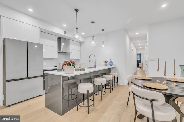 kitchen with wall chimney exhaust hood, light hardwood / wood-style flooring, hanging light fixtures, fridge, and white cabinets