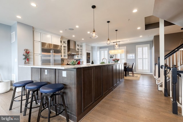 kitchen with wall chimney exhaust hood, a breakfast bar area, white cabinetry, built in fridge, and decorative backsplash