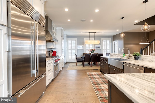 kitchen with sink, high end appliances, white cabinetry, hanging light fixtures, and wall chimney range hood